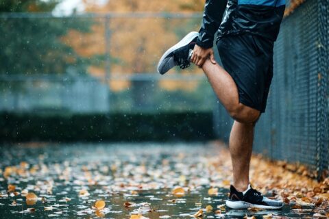 Lower section of a man stretching his quad in a tennis court on a rainy fall day