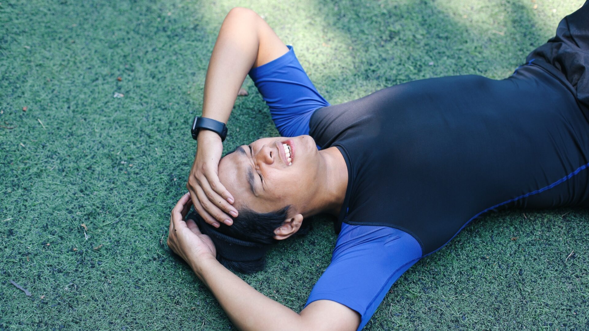 An athlete lies on grassy turf, holding his head and grimacing.