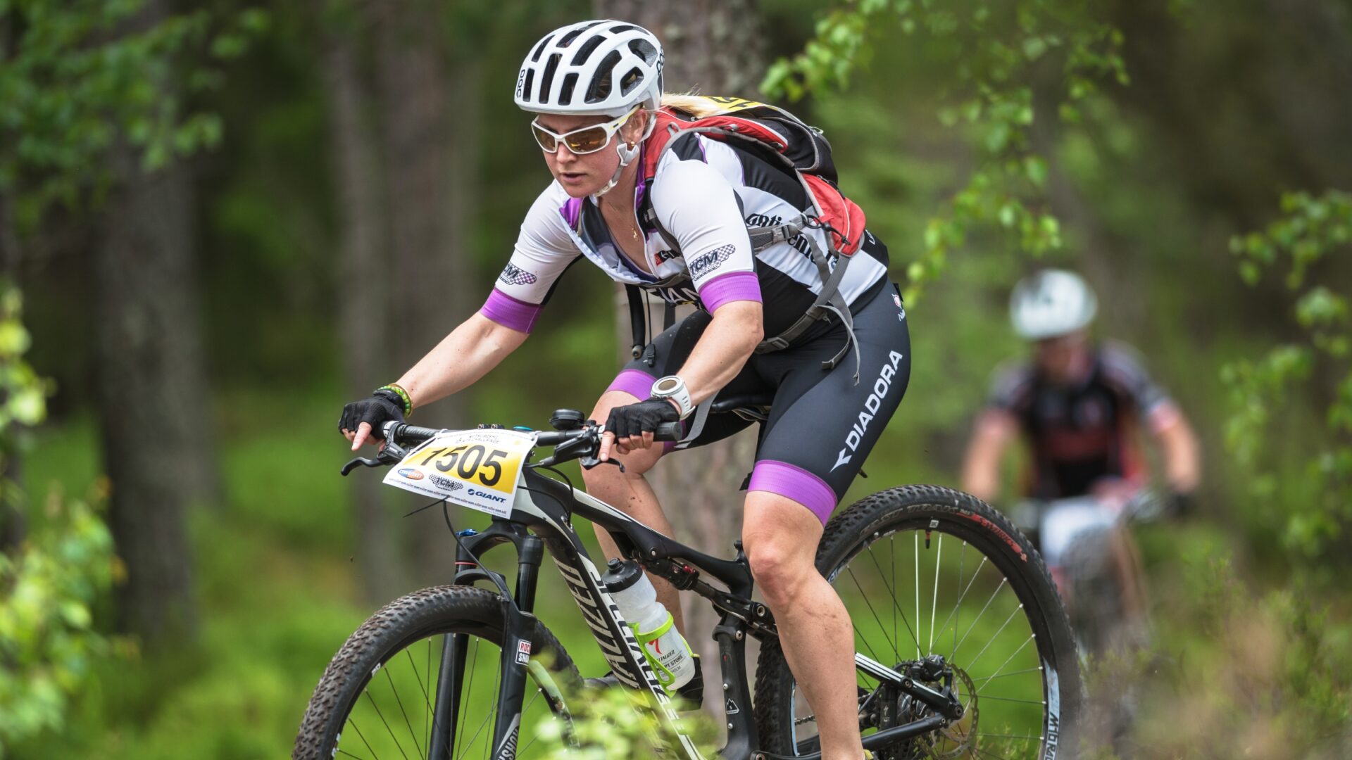 A female cyclist rides through a green forest on a mountain bike. She is wearing a hydration pack and has a water bottle on her bike.