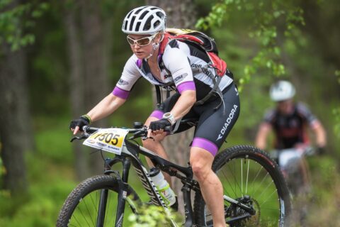 A female cyclist rides through a green forest on a mountain bike. She is wearing a hydration pack and has a water bottle on her bike.