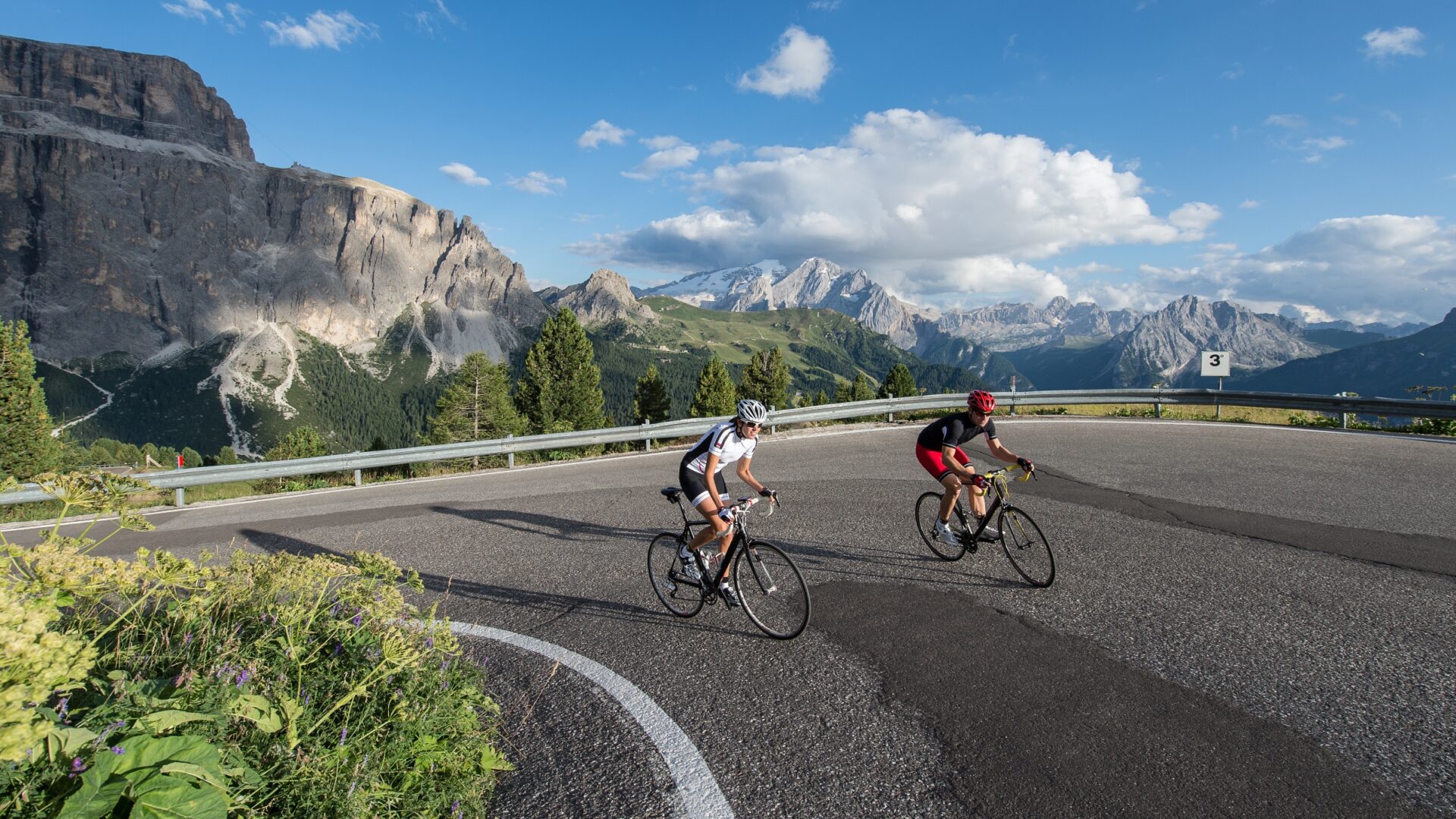 Two cyclists round the turn of a mountain climb on a sunny day